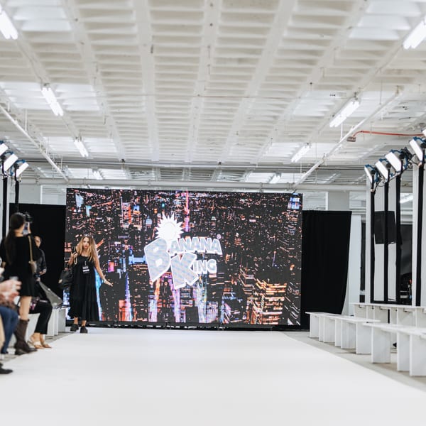 A woman in a black dress walks a runway with the Banana King logo on a large screen.