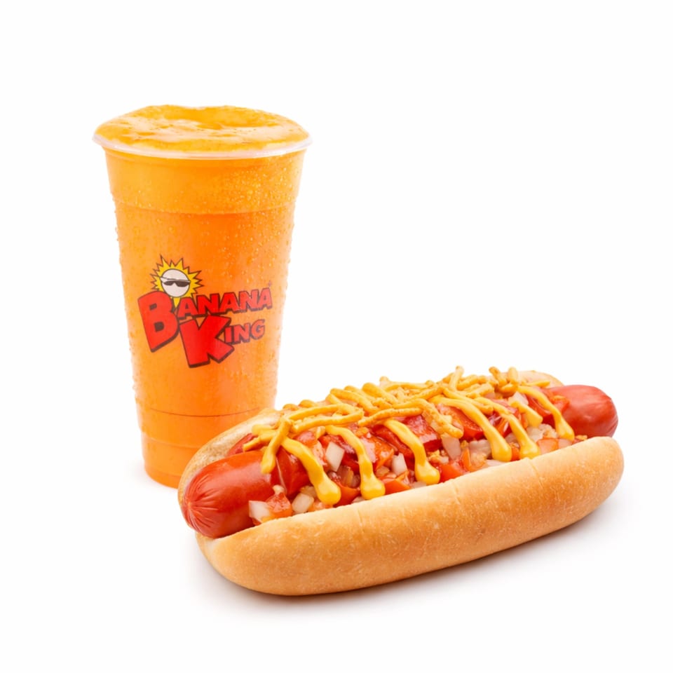 Hot dog topped with diced onions, tomatoes, shredded cheese, and yellow mustard beside an orange drink in a Banana King cup.