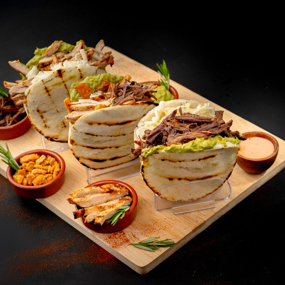 Three arepas filled with various meats, guacamole, and cheese, served on a wooden board.