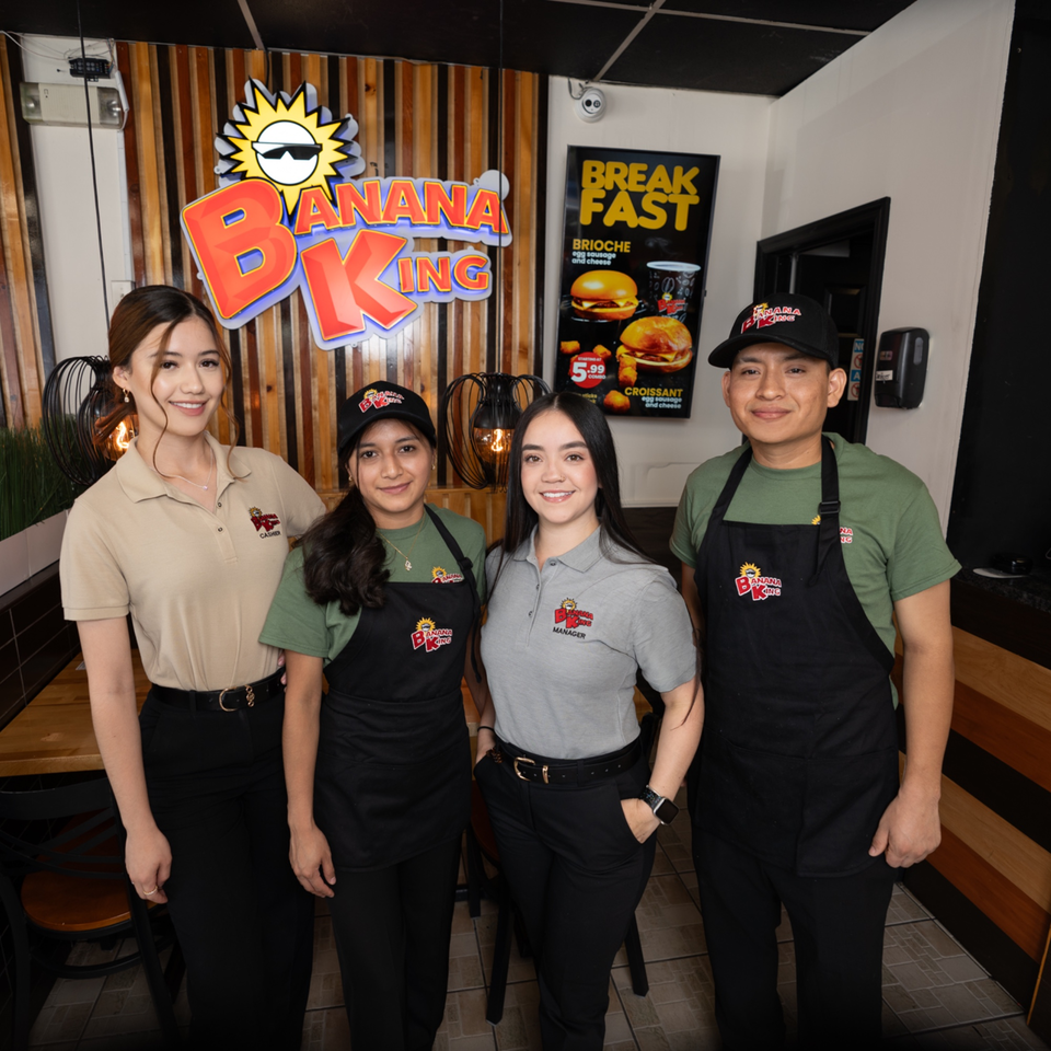 Four Banana King employees, two women and two men, stand smiling in front of the restaurant's logo and breakfast menu.