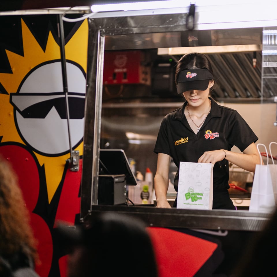 A Banana King employee in a food truck prepares an order in a branded paper bag.