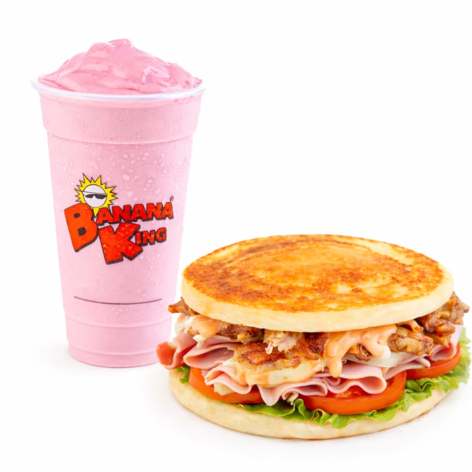 Banana King chicken sandwich with lettuce, tomatoes, ham, crispy chicken, and sauce beside a pink smoothie.